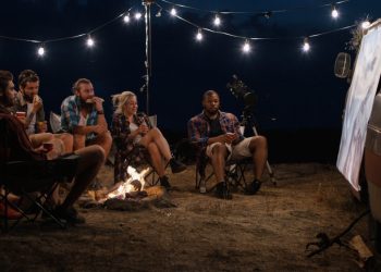 Amigos multiétnicos relajándose alrededor de una hoguera en el camping y viendo películas en la pantalla en la noche con guirnaldas brillantes encima