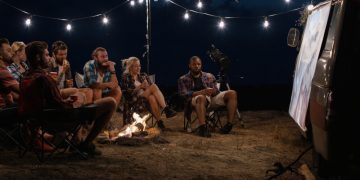 Amigos multiétnicos relajándose alrededor de una hoguera en el camping y viendo películas en la pantalla en la noche con guirnaldas brillantes encima