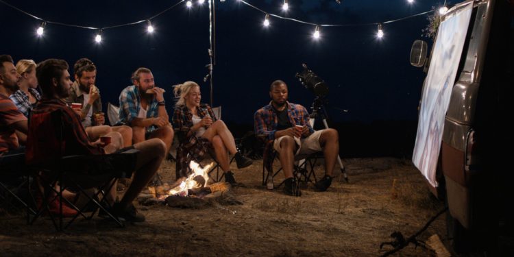 Amigos multiétnicos relajándose alrededor de una hoguera en el camping y viendo películas en la pantalla en la noche con guirnaldas brillantes encima