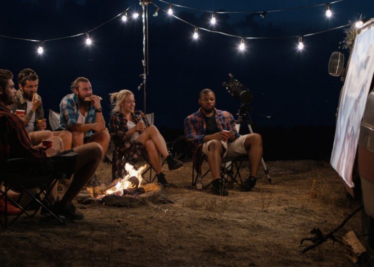 Amigos multiétnicos relajándose alrededor de una hoguera en el camping y viendo películas en la pantalla en la noche con guirnaldas brillantes encima
